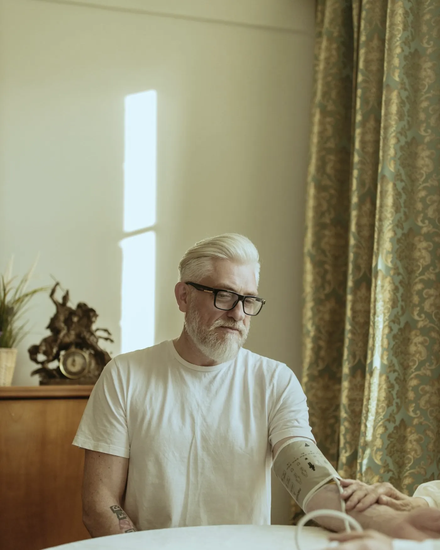 A gray-haired man in glasses having his blood pressure taken at a table at home