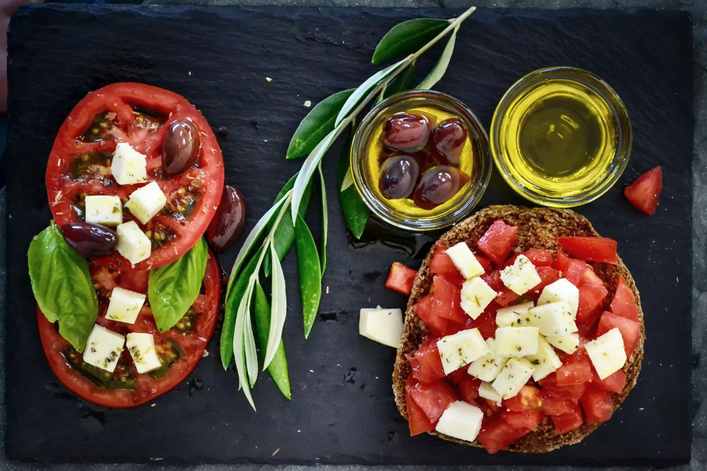 Tomatoes with feta and basil, olives, an olive branch and a small bowl of olive oil on a dark slate board