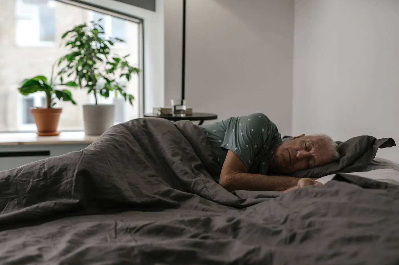 An older man asleep under a gray duvet in a bright, quiet bedroom
